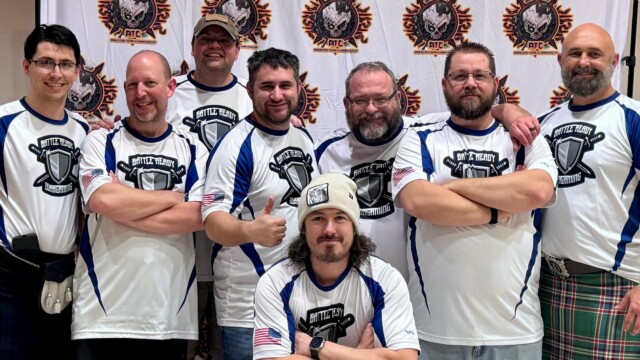 Eight men in matching white and blue Battle Ready jerseys pose together, smiling, in front of a backdrop with orange and black emblems. Showing their love for their gaming hobby, two on the sides wear kilts, while one in front sports a beanie. Eight men in matching white and blue Battle Ready jerseys pose together, smiling, in front of a backdrop with orange and black emblems. Showing their love for their gaming hobby, two on the sides wear kilts, while one in front sports a beanie.