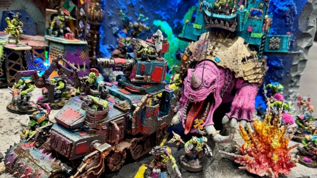 A detailed tabletop diorama for the gaming hobby features colorful orc miniatures, an armored vehicle with spikes and guns, a large pink dinosaur-like creature with orcs riding it, and vibrant neon-lit scenery—perfect for inspiring young builders like Phoebe Surgeon. Miniature orc figures battle with a spiked vehicle and purple dinosaur in a vibrant diorama, inspiring kids’ imaginations.