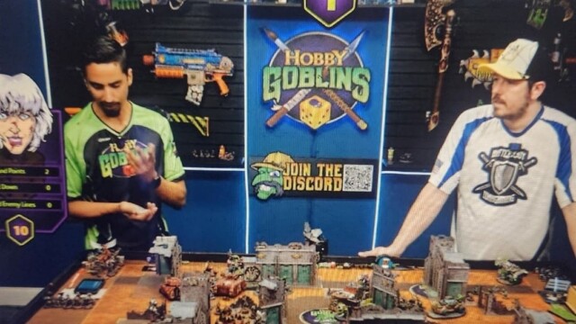Two men stand at a gaming table covered with miniature terrain and figures, deep in discussion about their gaming hobby. Behind them is a “Hobby Goblins” sign, game stats on the left, and a Discord QR code in the center. Two men stand at a gaming table covered with miniature terrain and figures, deep in discussion about their gaming hobby. Behind them is a “Hobby Goblins” sign, game stats on the left, and a Discord QR code in the center.