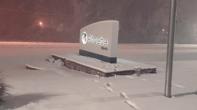 A snow-covered sign for Phoebe Worth stands outside at night, illuminated under streetlights after a winter storm. Fresh snow blankets the ground, with visible footprints leading toward the sign, signaling normal operations amid the wintry scene.