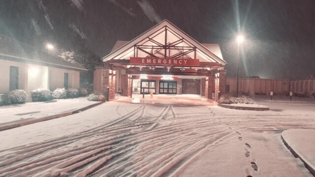 Snow covers the ground and drive leading to Phoebe’s emergency room entrance at night. Warm lights illuminate the building, while tire tracks and footprints in the fresh snow reflect normal operations during a winter storm.