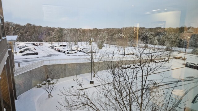 A snowy parking lot viewed from a building window, with parked cars, bare trees, and a snow-covered landscape. Despite the winter storm, normal operations continue as the calm and wintry scene unfolds under cloudy skies.