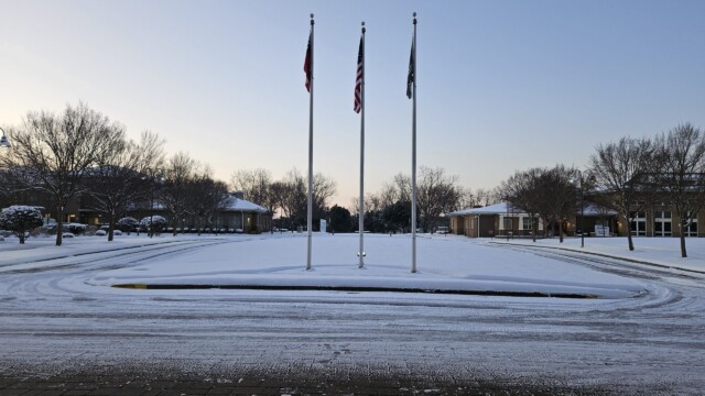 Three flagpoles stand in a snowy courtyard surrounded by buildings and bare trees under a clear sky. Despite the aftermath of a recent winter storm, the area appears quiet and empty, with tire tracks hinting at normal operations resuming.