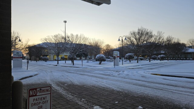 A snow-covered parking lot at sunrise after a winter storm, with tire tracks, bare trees, and a medical facility sign. A No Parking, Patient Drop-Off or Pick-Up Only sign is visible in the foreground as normal operations resume.