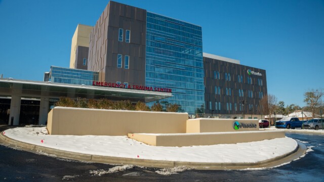 Modern hospital building with Emergency & Trauma Center sign at the entrance, surrounded by a snow-dusted landscape after a winter storm. The Phoebe hospital logo and name are visible, assuring normal operations under a clear blue sky.