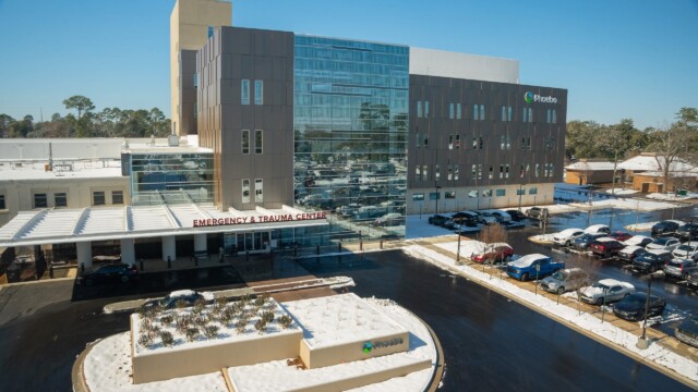The Phoebe Emergency & Trauma Center, with glass and brown paneling, continues normal operations amid a winter storm, surrounded by snow-covered ground and parked cars under a clear blue sky.