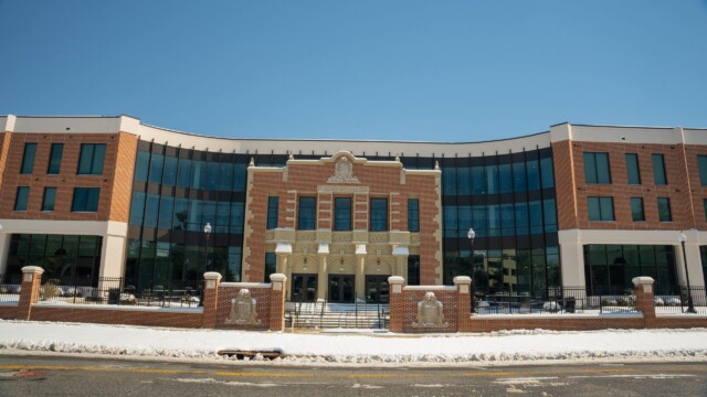 A modern brick and glass building with a central entrance, set behind a low brick fence. After a winter storm, snow covers the ground beneath clear skies. The building blends both historic and contemporary architectural elements. Normal operations have resumed.