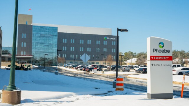 A hospital building with Phoebe signage stands on a snowy day after a winter storm. In the foreground, a sign points to Emergency and Labor & Delivery as cars are parked nearby and snow blankets the ground, indicating normal operations continue.
