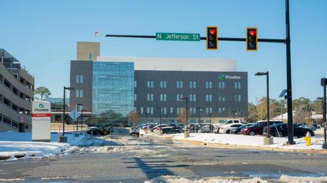 A snow-covered street leads to a modern hospital building with Phoebe signage, beneath a street sign for N Jefferson St and a red traffic light, as normal operations continue despite the recent winter storm on a clear, sunny day.