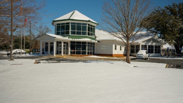 A modern building with large windows and a green roof sits surrounded by snow left from a recent winter storm, with bare trees and a parked car nearby under a clear blue sky as Phoebe ensures normal operations continue smoothly.