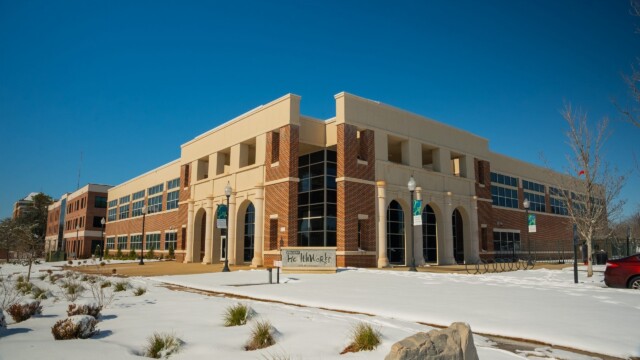 A large brick and tan building with arched windows sits under a clear blue sky. Despite a recent winter storm, snow covers the ground with patches of grass peeking through. A sign and lampposts are visible in front as Phoebe resumes normal operations.