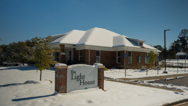 A brick building with a sign reading The Light House stands under a clear sky, surrounded by snow-covered ground and trees after a winter storm. Accessible parking signs are visible in the nearly empty lot, signaling normal operations.