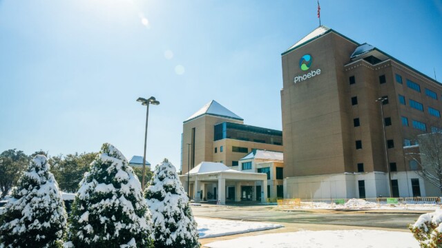 A hospital building with a Phoebe sign stands under a clear blue sky, surrounded by snow-covered trees and ground after a winter storm, with sunlight creating a bright and crisp winter scene as normal operations continue.