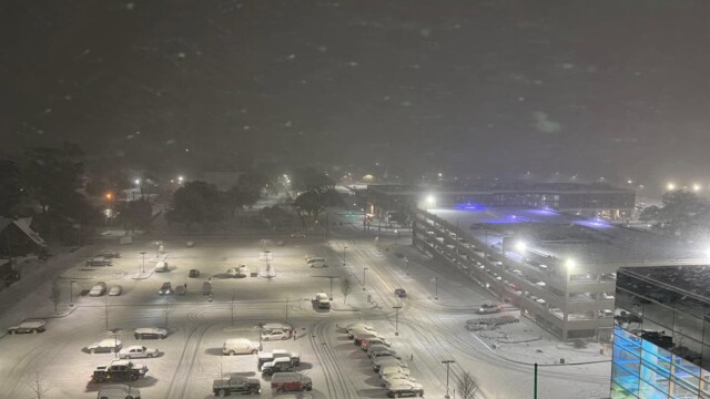 A nighttime view of a snow-covered parking lot and nearby garage during a winter storm, with cars parked and tire tracks visible. Streetlights and building lights create a soft glow through falling snow, suggesting normal operations continue for Phoebe.
