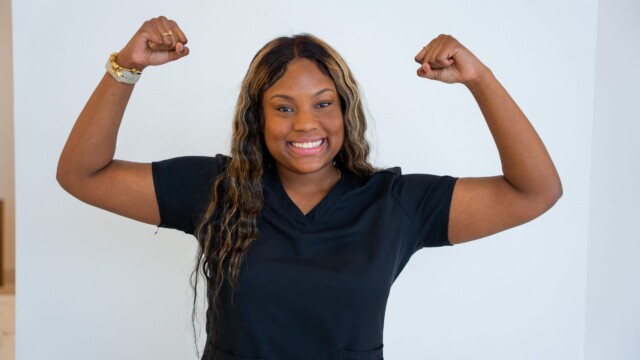 A woman wearing a black shirt smiles confidently while raising both arms and showing her biceps in a flexing pose, embodying the strength of a dedicated NICU Nurse, against a plain white background.