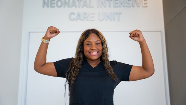 A smiling NICU nurse in scrubs stands before a Neonatal Intensive Care Unit sign, arms raised in a strong, triumphant pose.