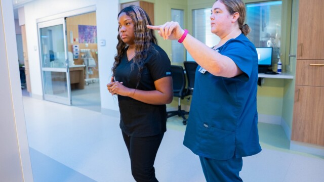 Two healthcare professionals in scrubs walk through a modern hospital, reflecting an inspiring journey. One, possibly a NICU nurse like Hannah Lockett, points ahead to give guidance while the other listens attentively.