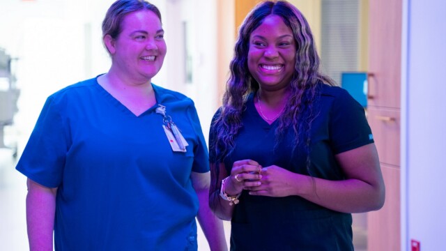 Two women in medical scrubs stand and smile together in a brightly lit hallway—one wearing a name badge, the other holding her hands—a snapshot of an inspiring journey, perhaps between a NICU nurse and someone touched by her care.