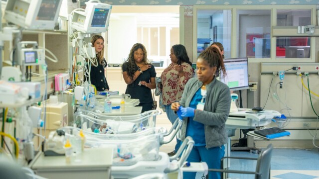 A hospital room with medical equipment and empty baby bassinets. Four women are talking in the background, while a dedicated NICU nurse in blue scrubs and gloves stands in the foreground near the bassinets.